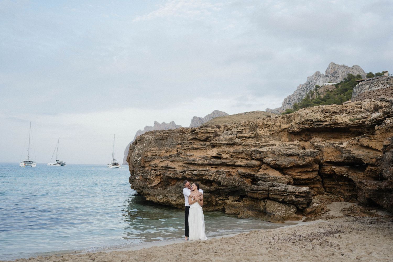Bride and groom embrace on a sandy beach beside a rocky cliff, with calm blue sea and sailboats anchored in the distance.