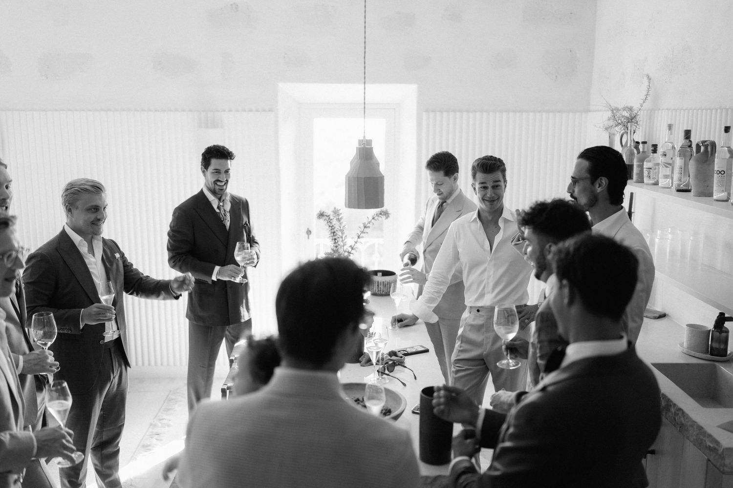 Group of men in smart attire raise glasses and chat around a bright kitchen counter during a lively wine gathering.