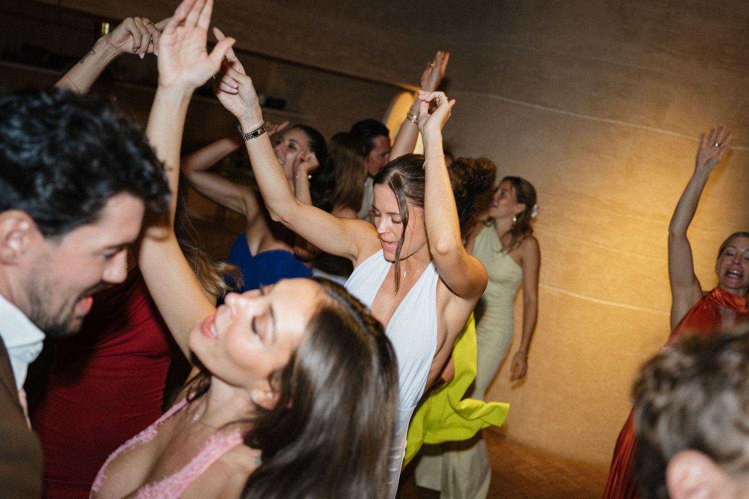 Guests dance joyfully with raised arms at a lively wedding reception as the bride in white smiles in the center.