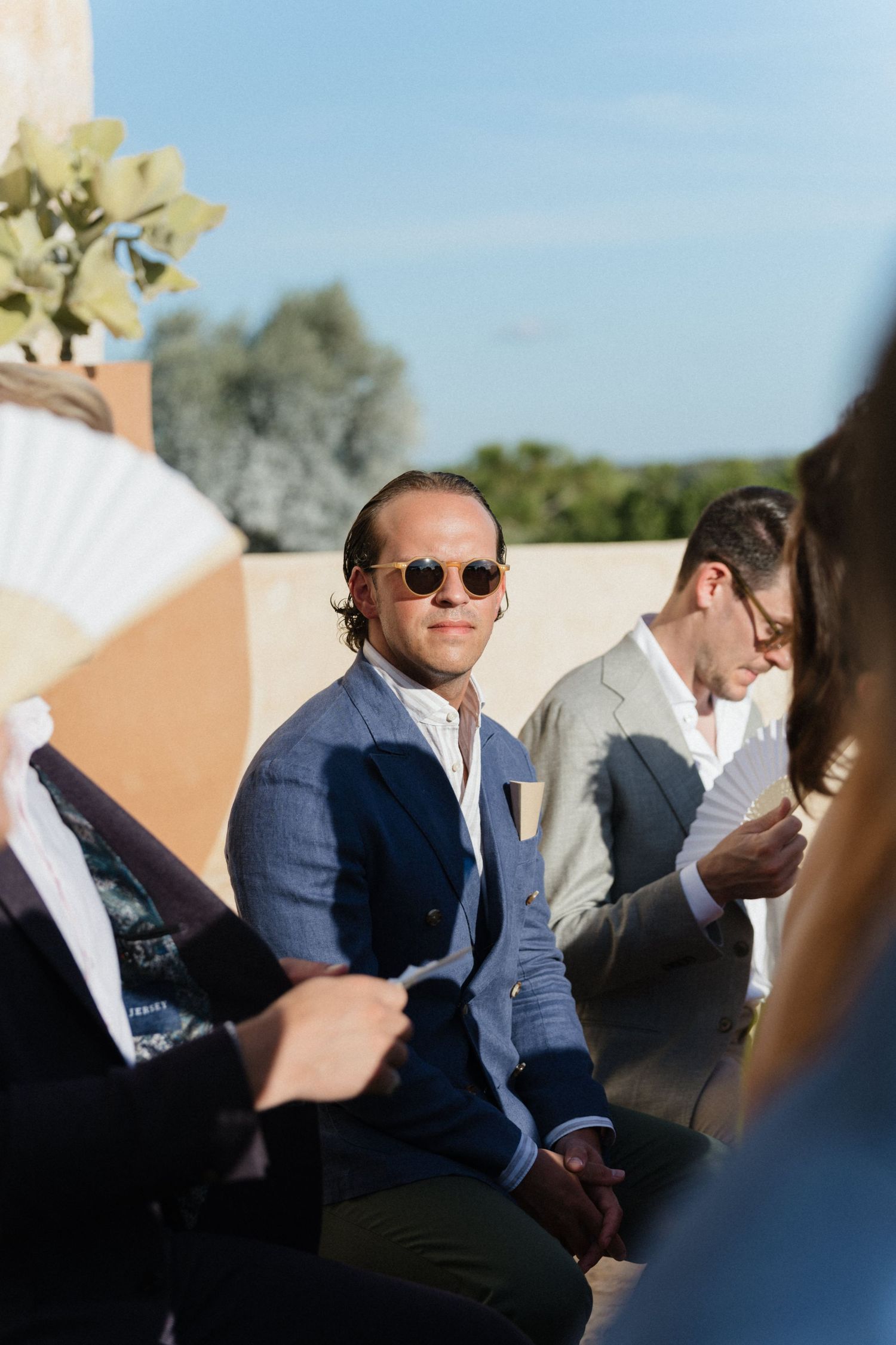 Man in round sunglasses and a blue blazer sits among guests at an outdoor wedding ceremony, with a hand fan in the foreground.