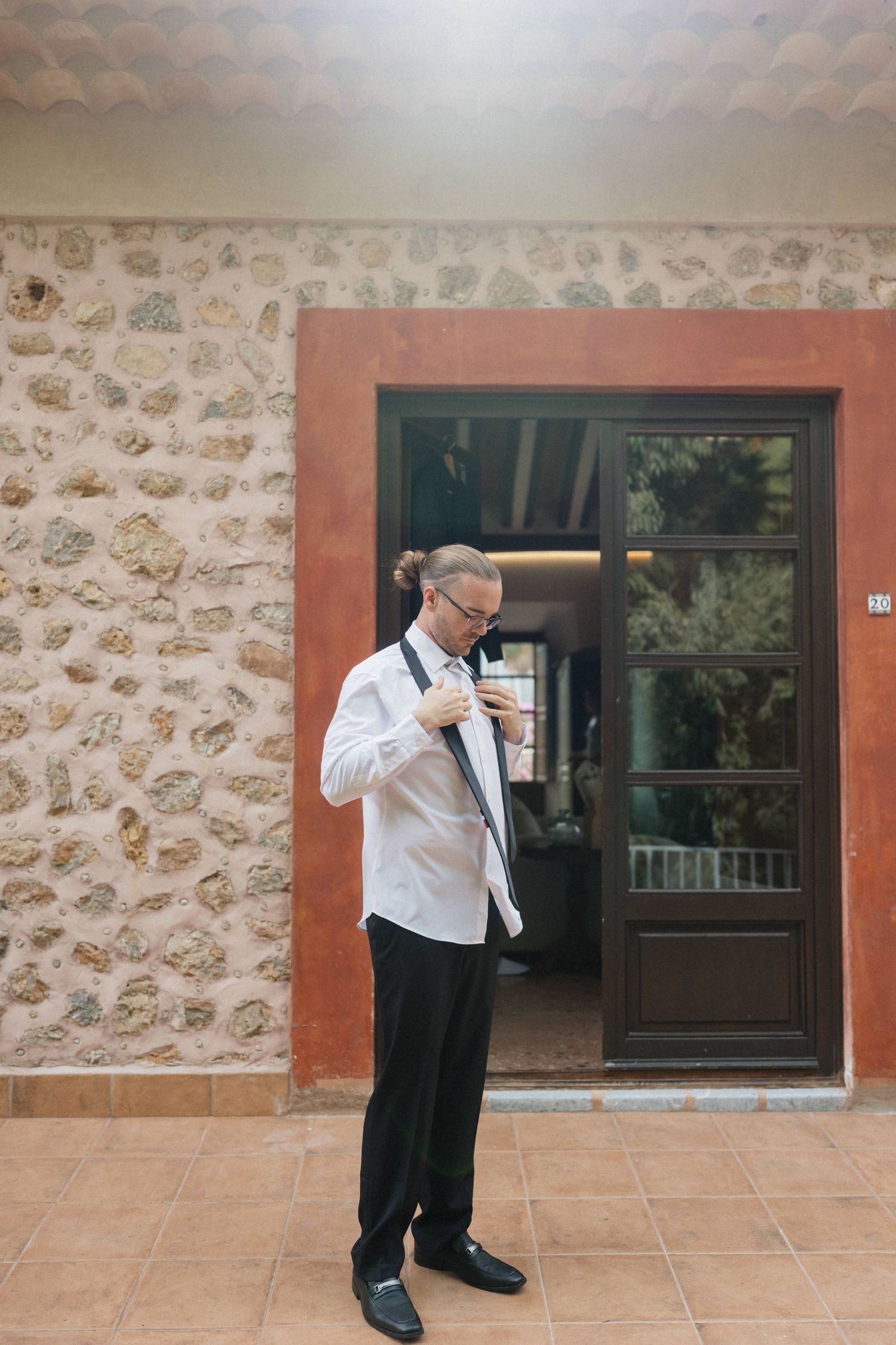 Man in a white dress shirt and black trousers adjusts his bow tie outside a rustic stone building with an orange door frame.