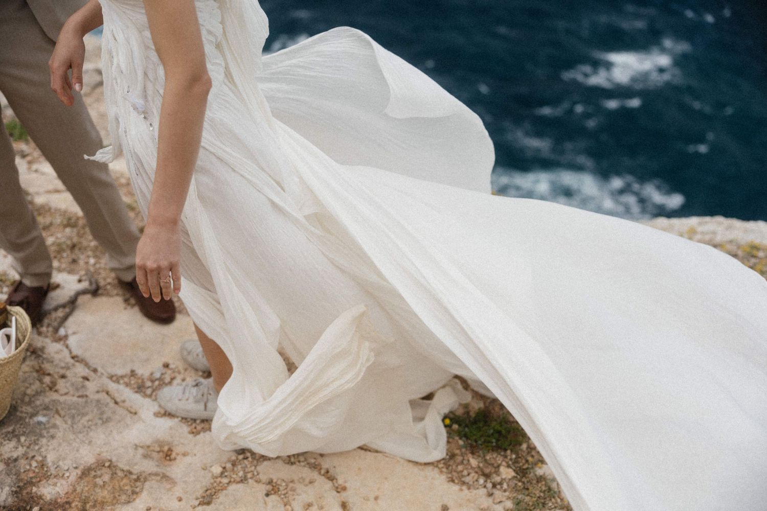 Bride in flowing white wedding dress standing on rocky cliff edge overlooking deep blue ocean waters.
