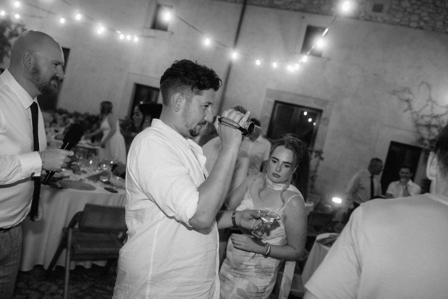 Black and white wedding reception photo of guests drinking and celebrating under string lights in an elegant venue.