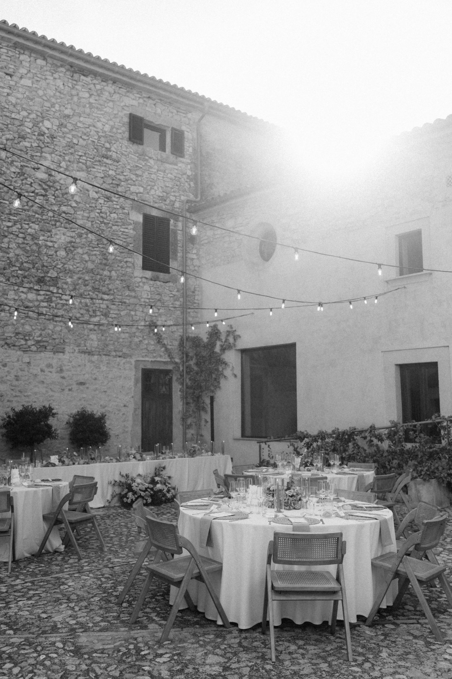 Black and white photo of outdoor wedding reception tables in a stone courtyard with string lights.