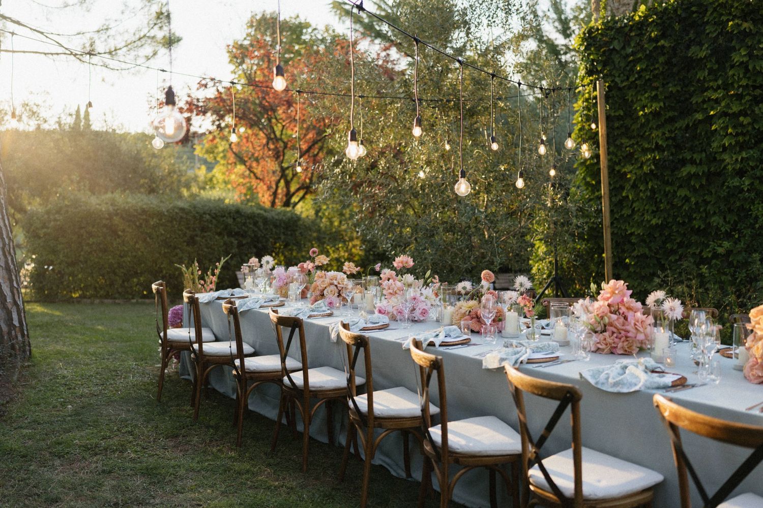 Romantic outdoor wedding reception table with pink floral arrangements and warm string lights at golden hour.