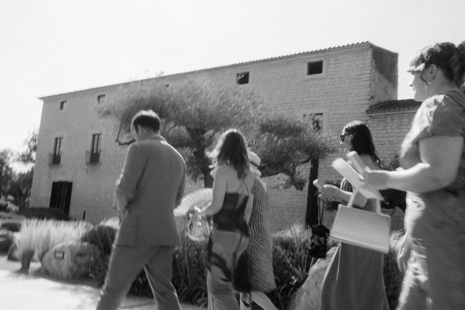 Group of people walking toward a rustic stone building surrounded by trees in a black and white outdoor photo.