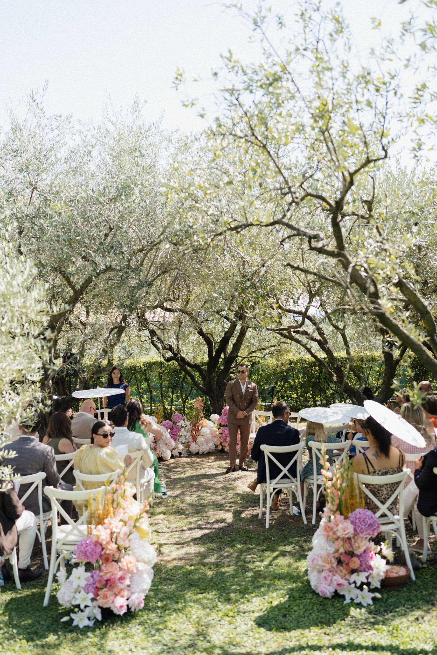 Outdoor wedding ceremony under olive trees with white chairs, pink floral arrangements, and guests enjoying a sunny garden setting.