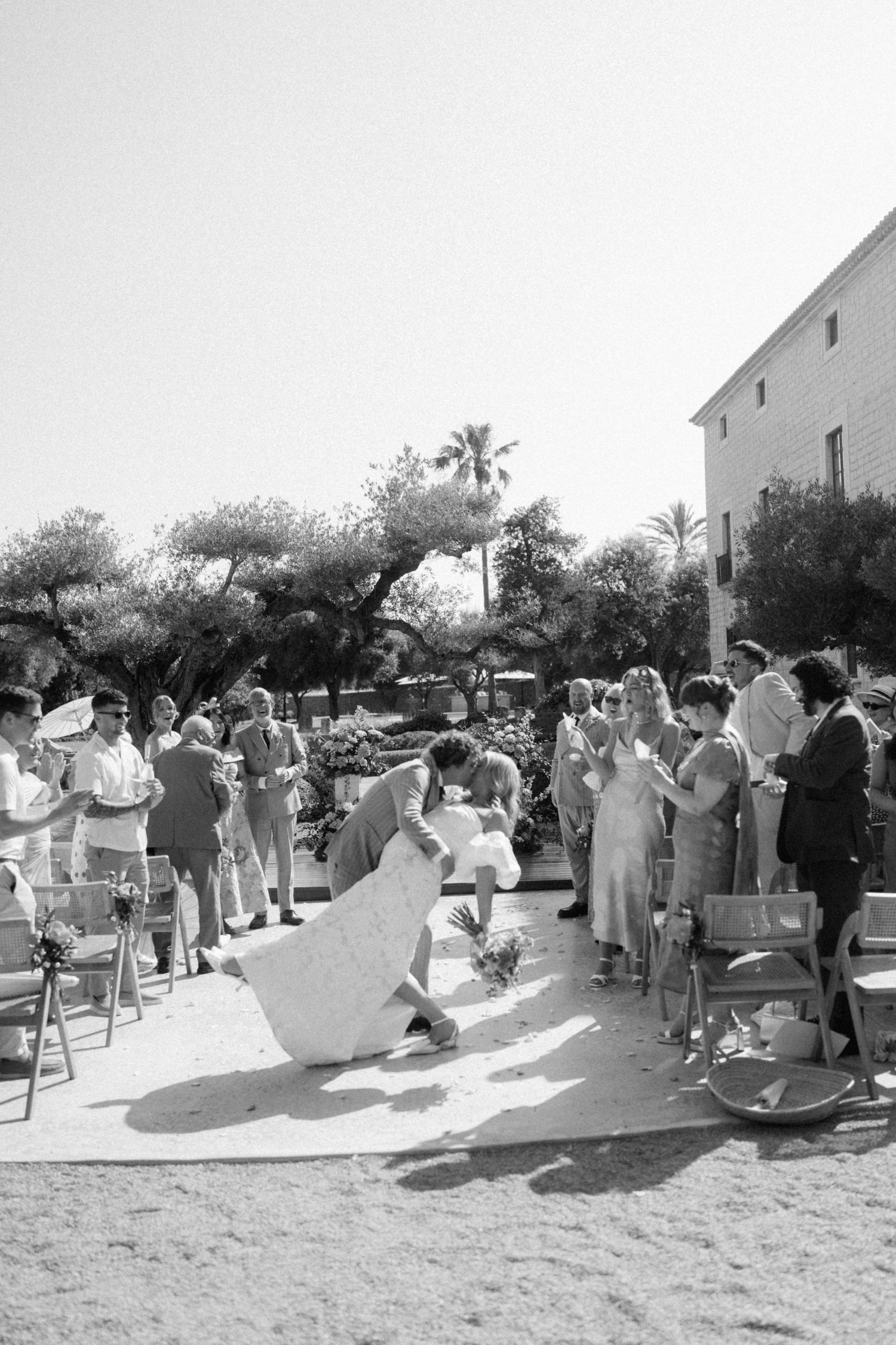 Black and white outdoor wedding ceremony with bride and groom walking down the aisle surrounded by guests and trees.