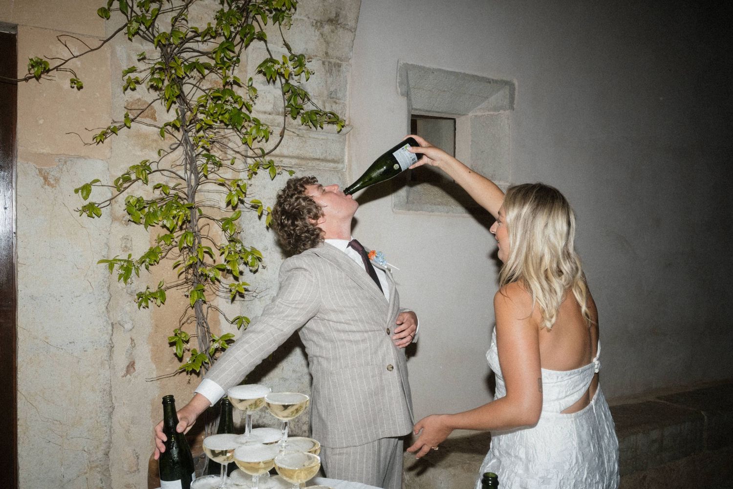 Bride pours champagne into groom's mouth from bottle at wedding reception near elegant table with glasses.