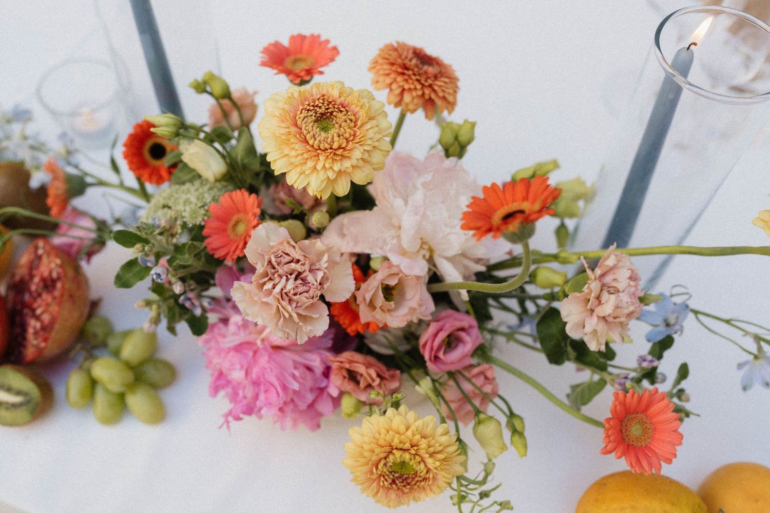 Colorful floral centerpiece with orange gerberas, pink carnations, and dahlias surrounded by grapes and citrus fruits on a white table.