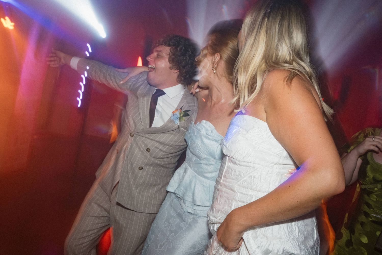 Three people dancing energetically at a wedding reception under colorful lights in a dimly lit venue.