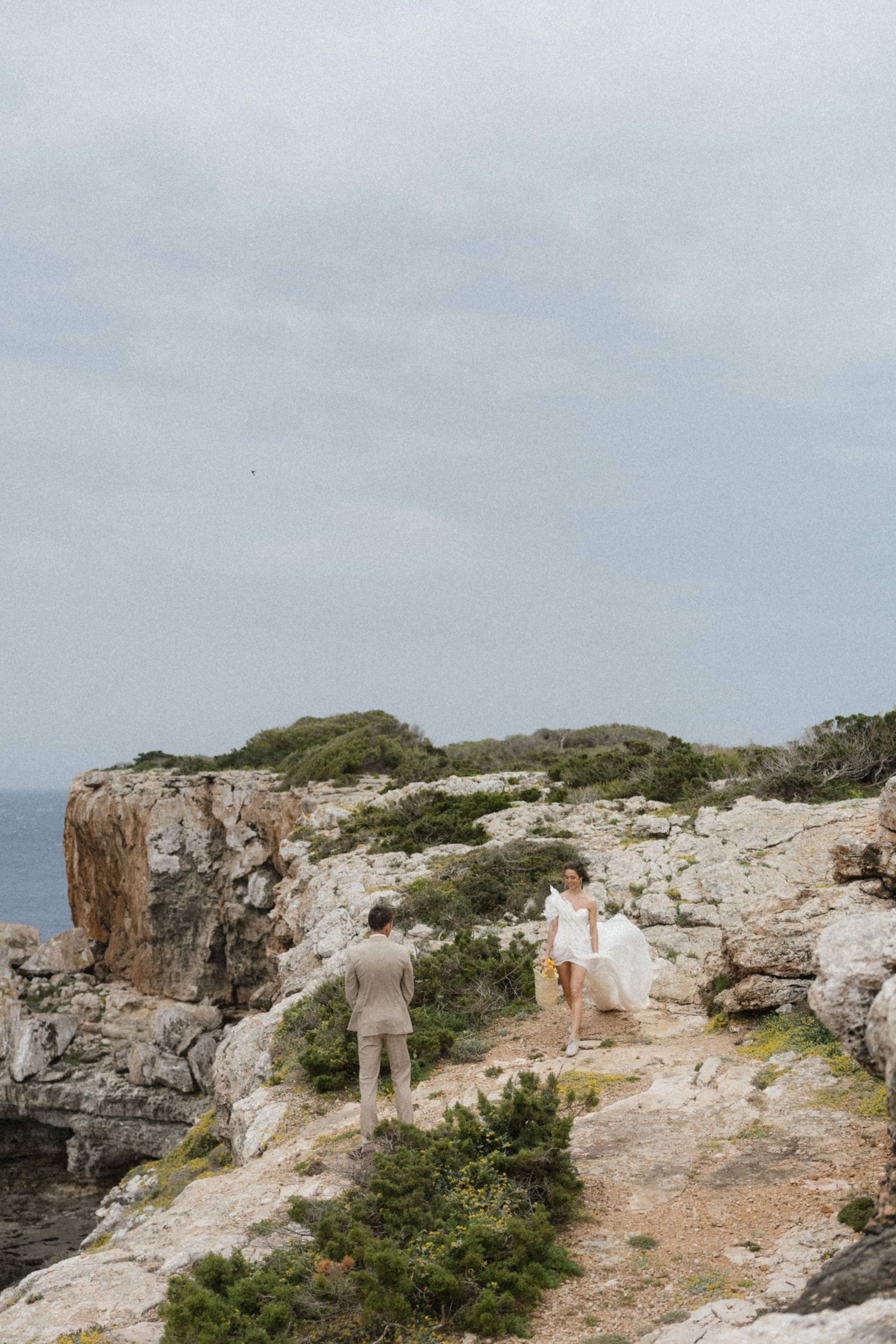Bride and groom walking on rocky coastal cliffs with dramatic ocean views on a cloudy day.
