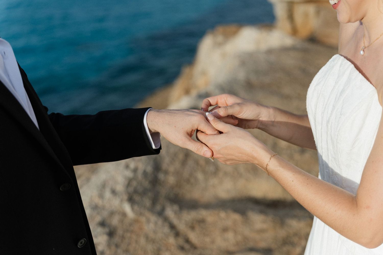 Groom in a dark suit places a wedding ring on the bride's finger beside the sea during a seaside ceremony.
