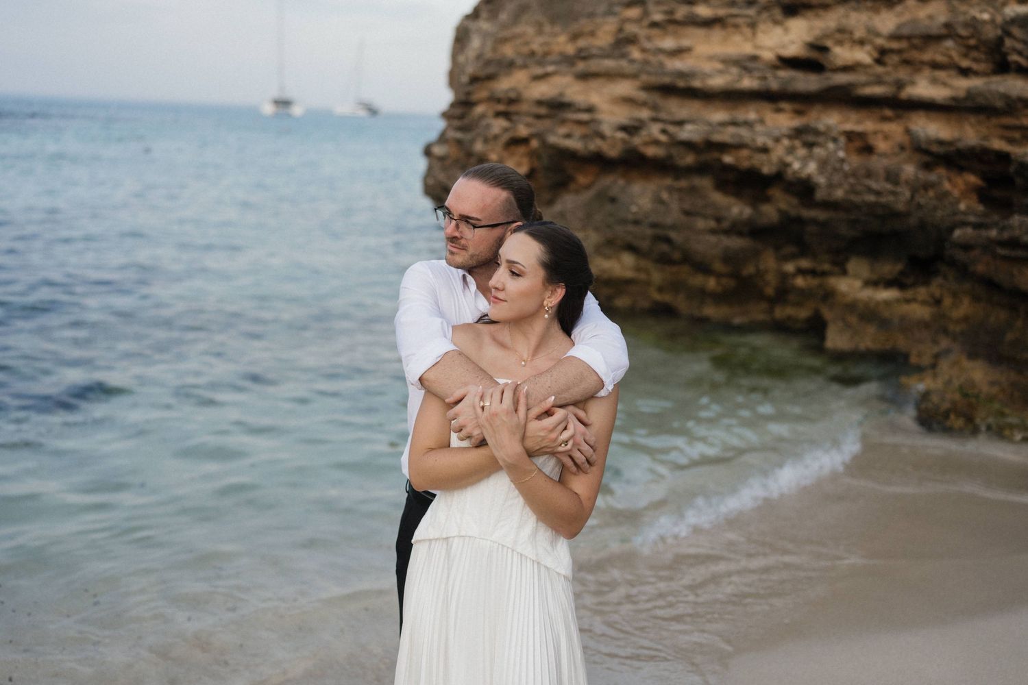 Couple embracing on a sandy beach beside turquoise water and rocky cliffs, with sailboats on the horizon, in a romantic seaside portrait.