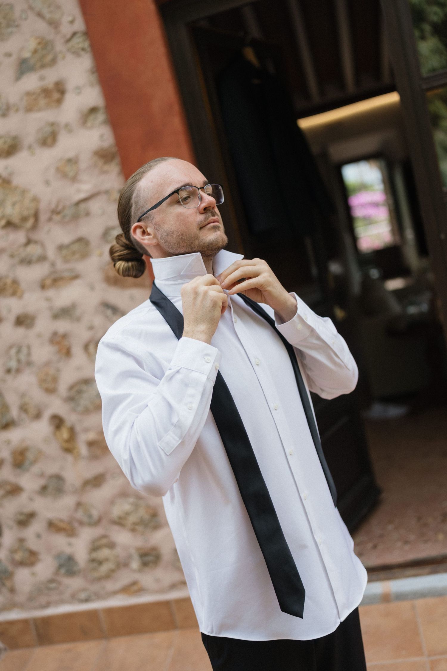 Man with glasses and a bun adjusts his collar, wearing a white dress shirt and loose black bow tie while getting ready outdoors.