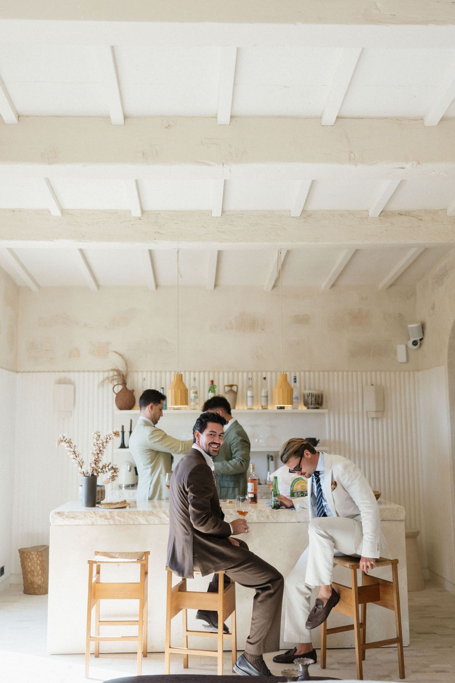 Three men in suits gather around a white marble bar in a bright rustic room with white wood beams, shelves of bottles and wooden stools.