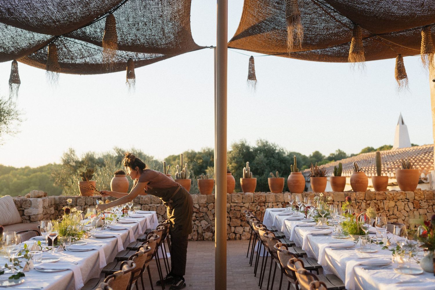 Staff member arranges a long outdoor wedding dinner table under a woven canopy with terracotta pots and olive trees.