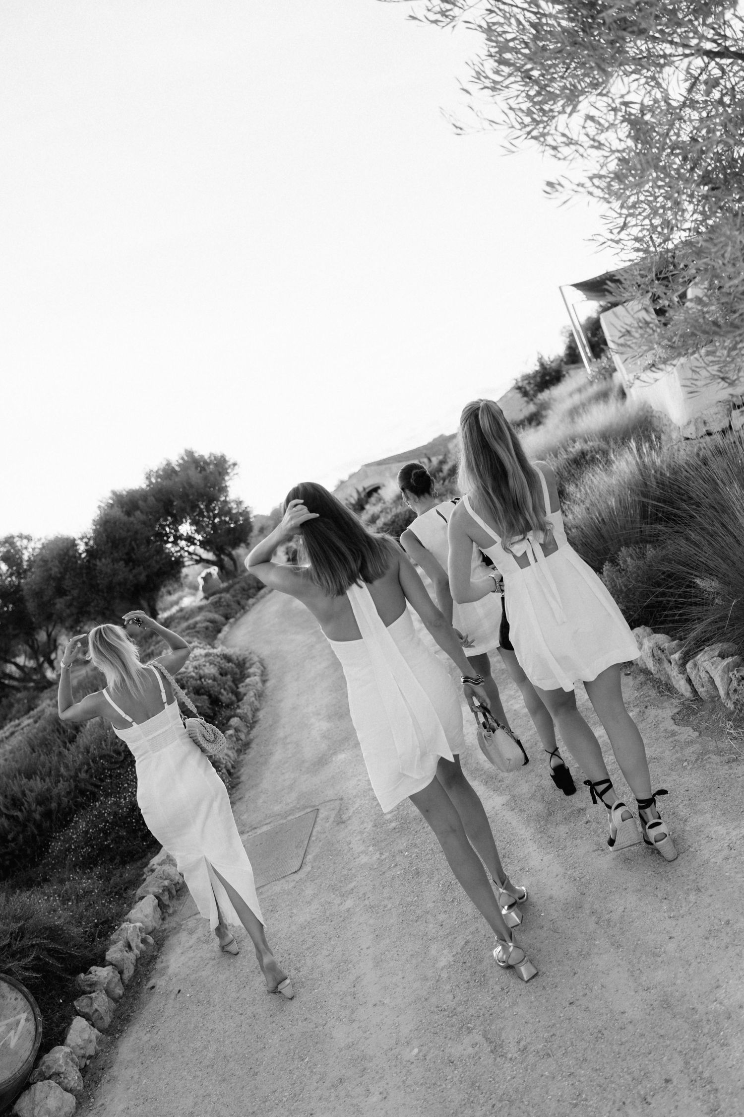Black and white photo of four women in white dresses walking away along a winding dirt path lined with shrubs and olive trees.