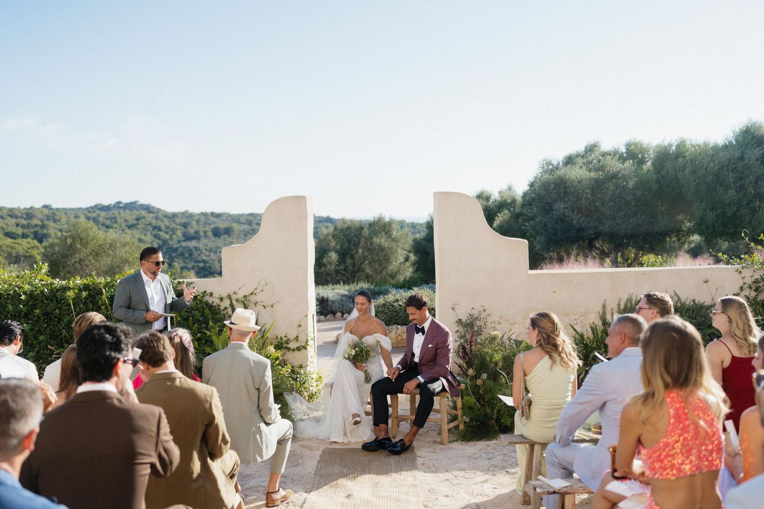Wedding guests sit in rows watching an outdoor ceremony as the bride and groom face the officiant between whitewashed stone walls.