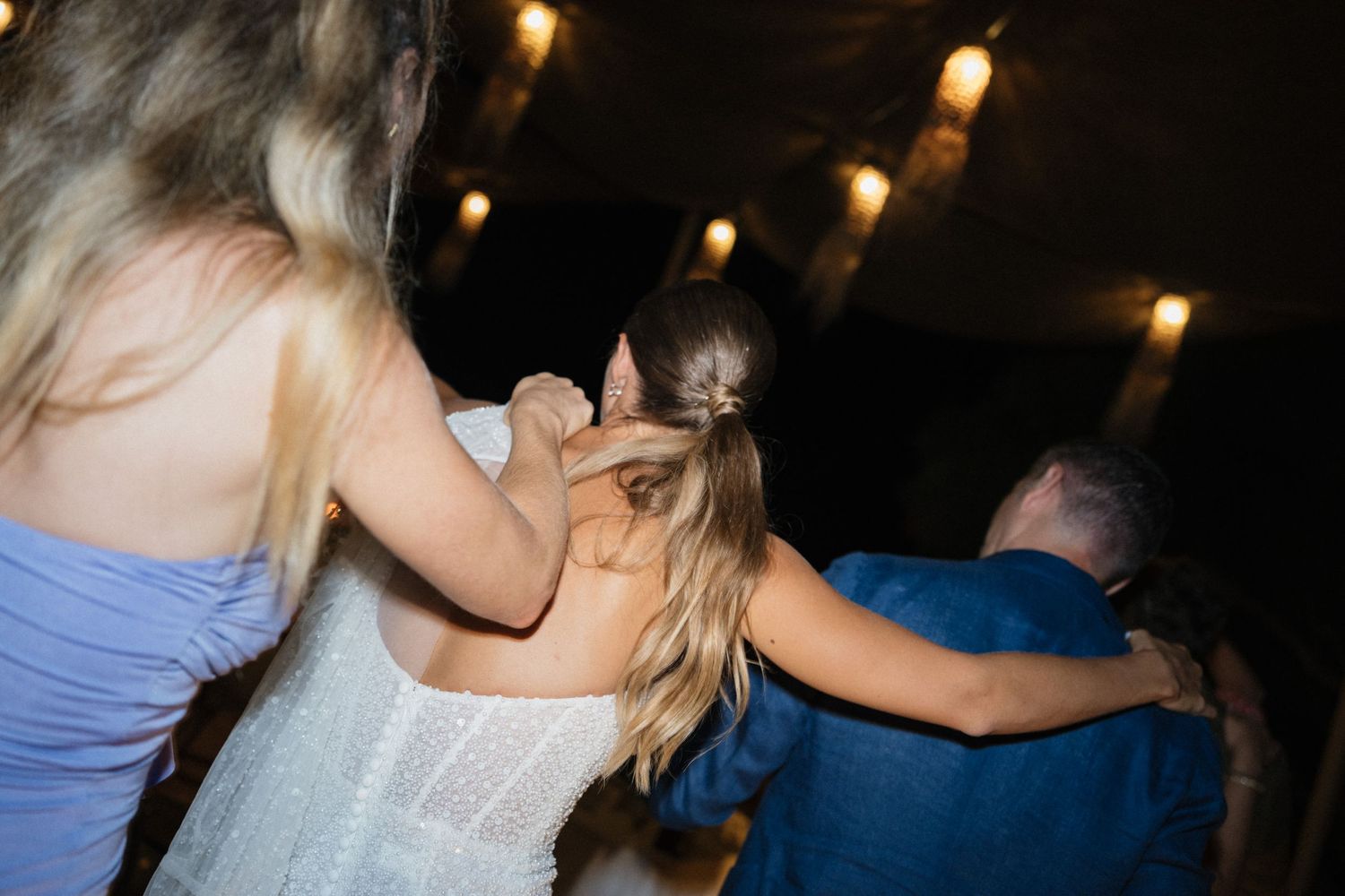 Wedding guests in blue and white dresses link arms and dance together under warm string lights at an evening celebration.