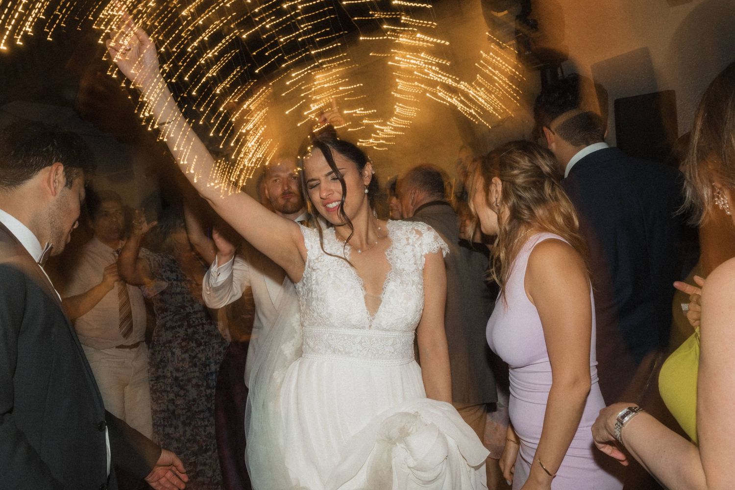Bride in a lace wedding gown dancing joyfully under sparkling fairy lights at a wedding reception.