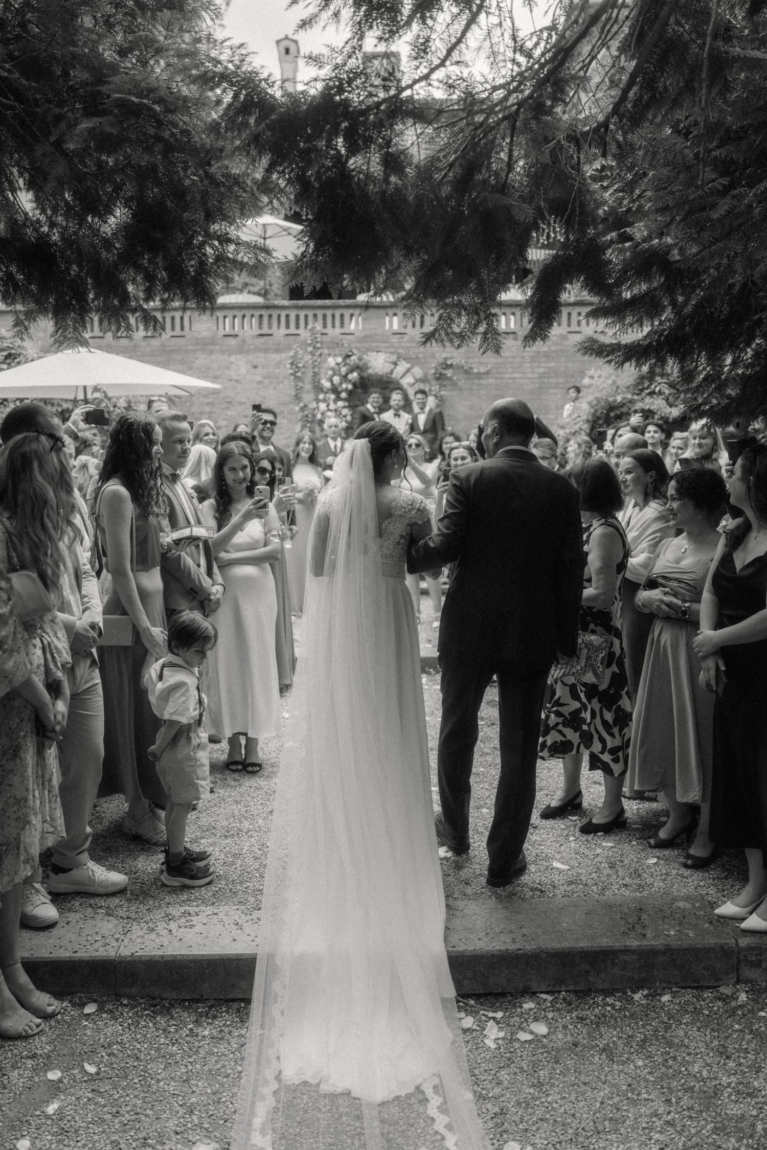 Black and white photo of a bride walking down the aisle with guests lining the path at an outdoor wedding ceremony.