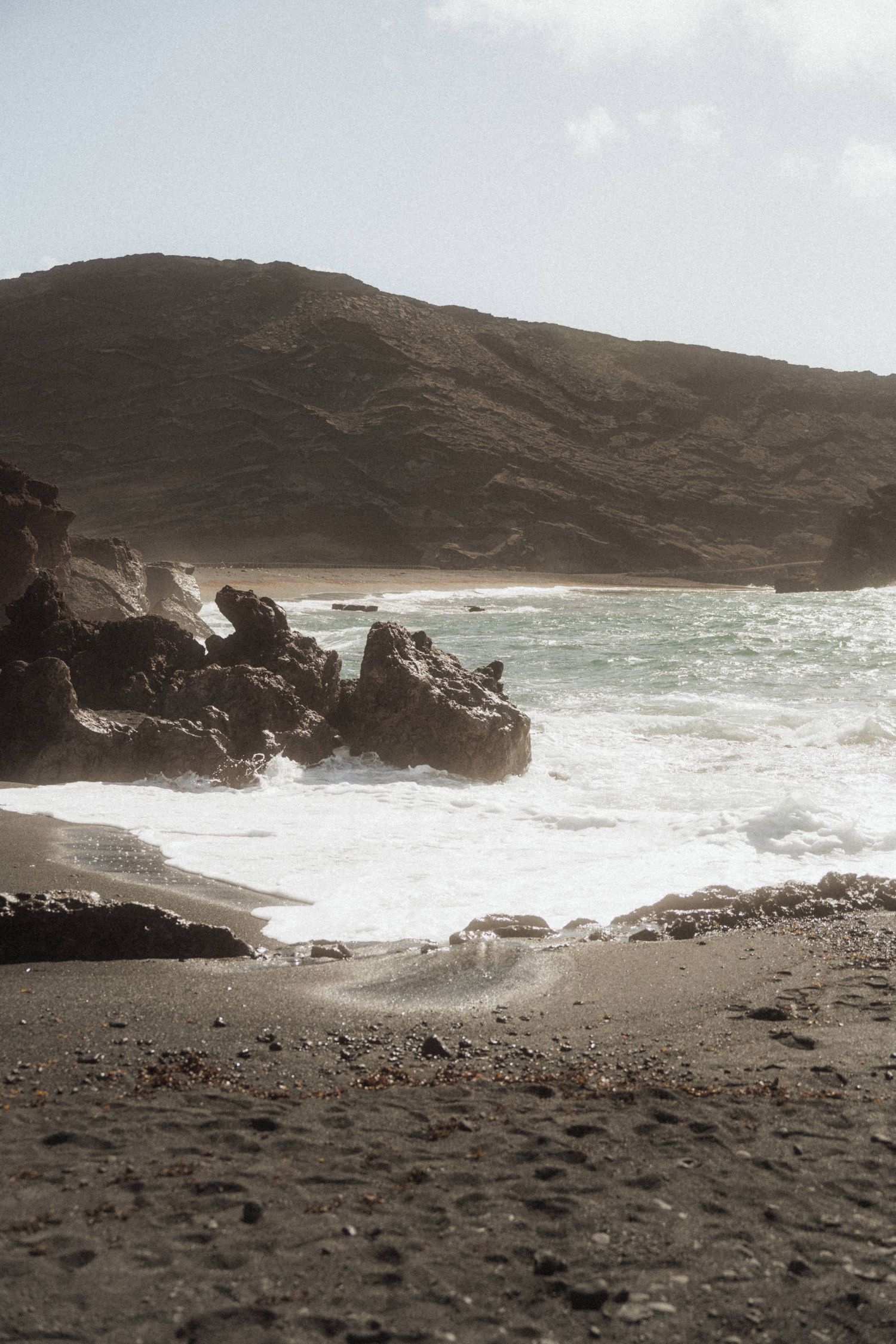 Dramatic volcanic black sand beach with crashing waves and rugged rocky coastline under a hazy sky.