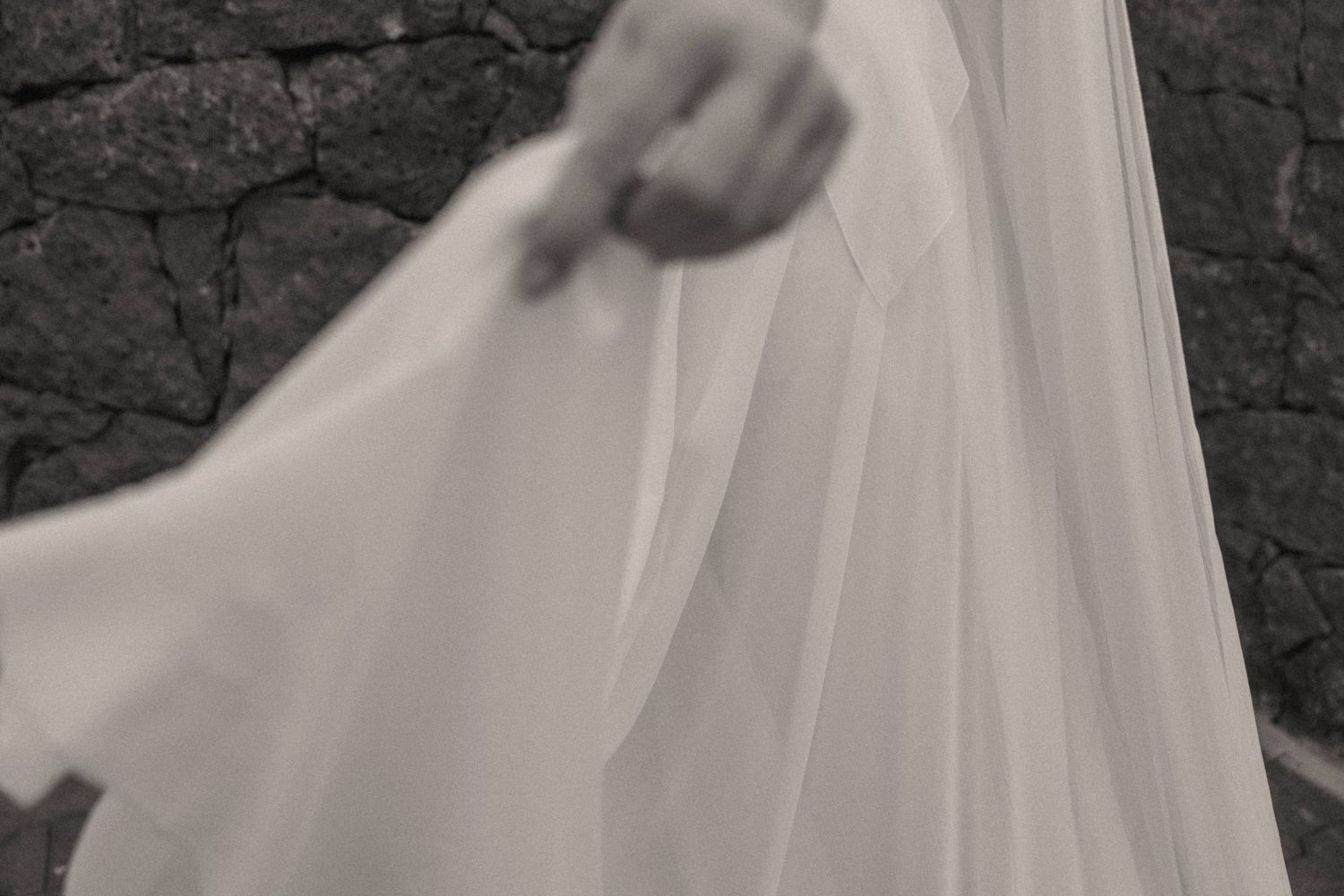Black and white photo of a bride holding her flowing white wedding veil against a dark stone wall.