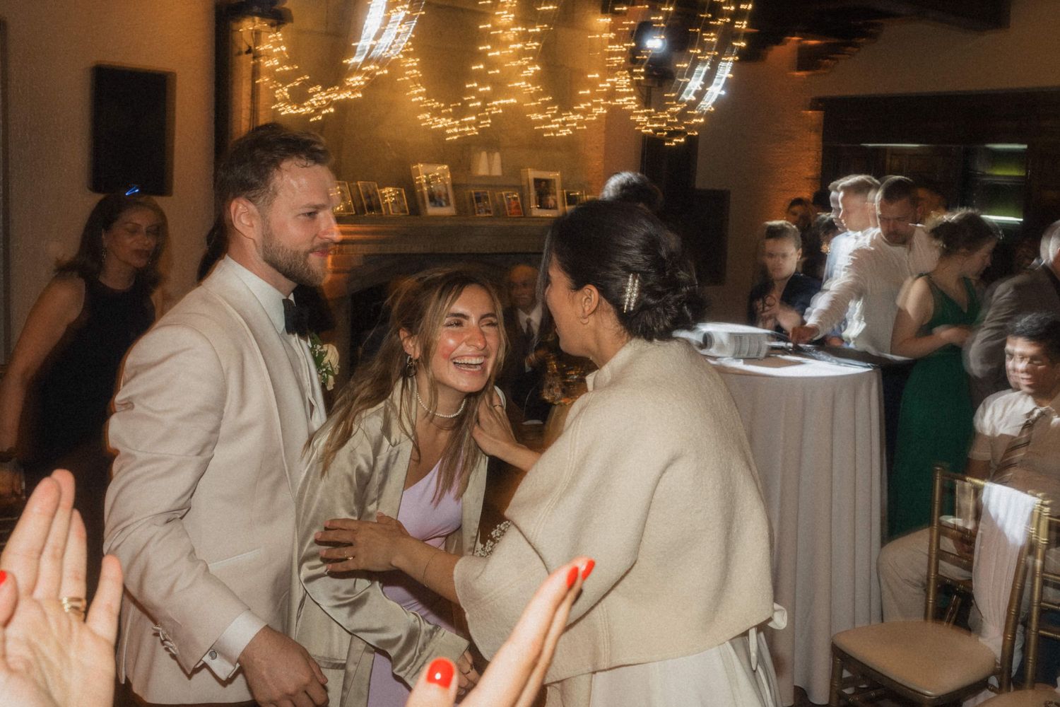 Guests laugh and embrace at a warmly lit wedding reception with fairy lights and decorations.