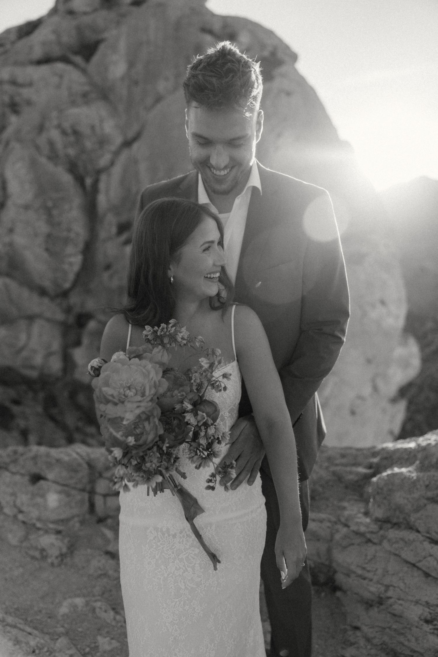 Black and white portrait of smiling couple embracing at sunset, bride in lace gown holding floral bouquet by rocks.