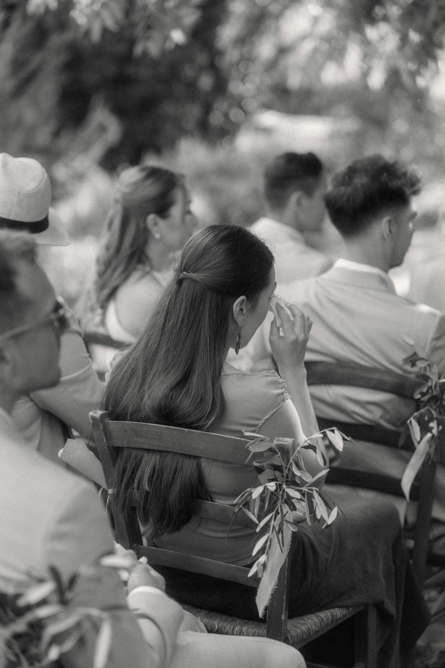 Black and white photo of wedding guests seated outdoors, a woman wiping tears during the ceremony.
