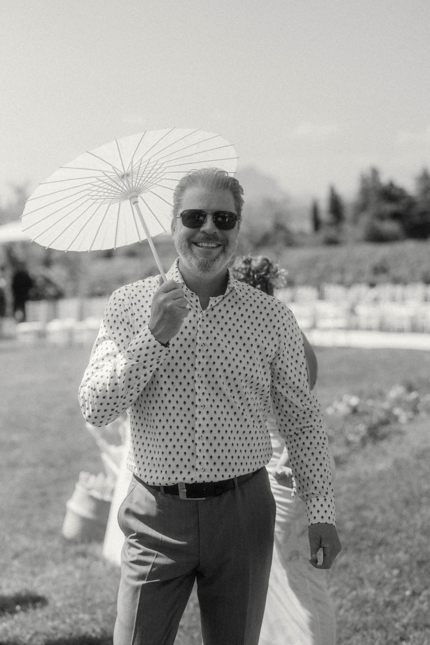 Smiling man in polka dot shirt holding white parasol umbrella at outdoor wedding ceremony.