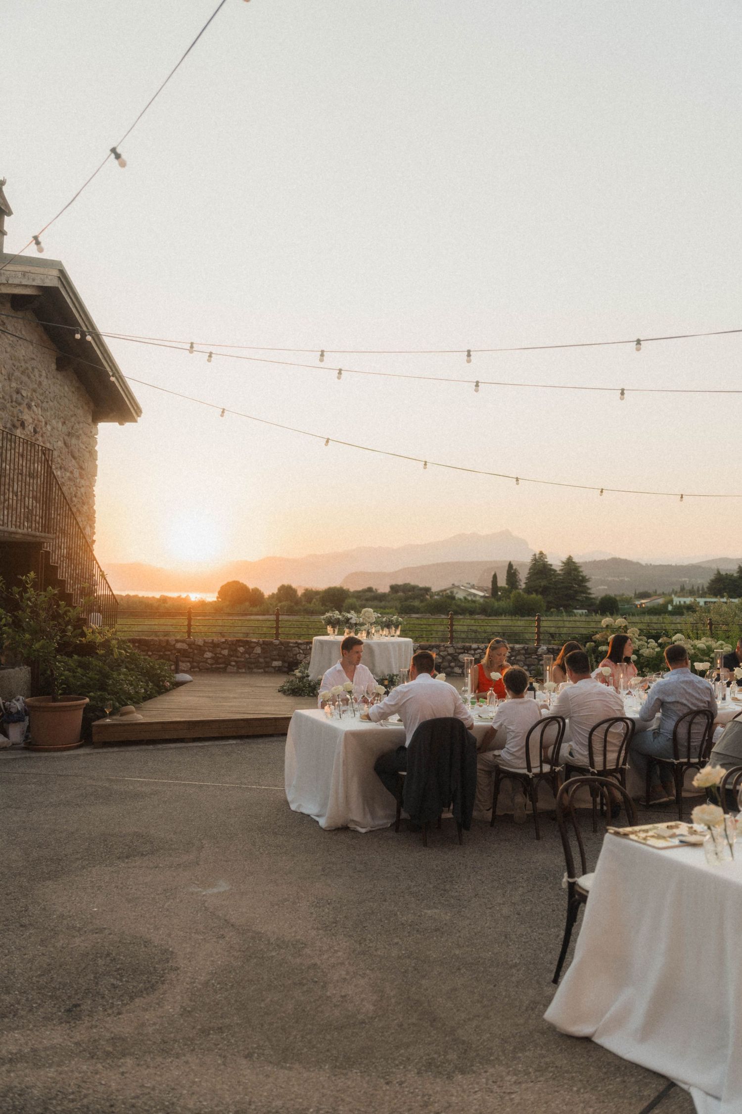 Wedding guests dining outdoors at sunset with string lights, mountain views, and golden hour glow over vineyard.