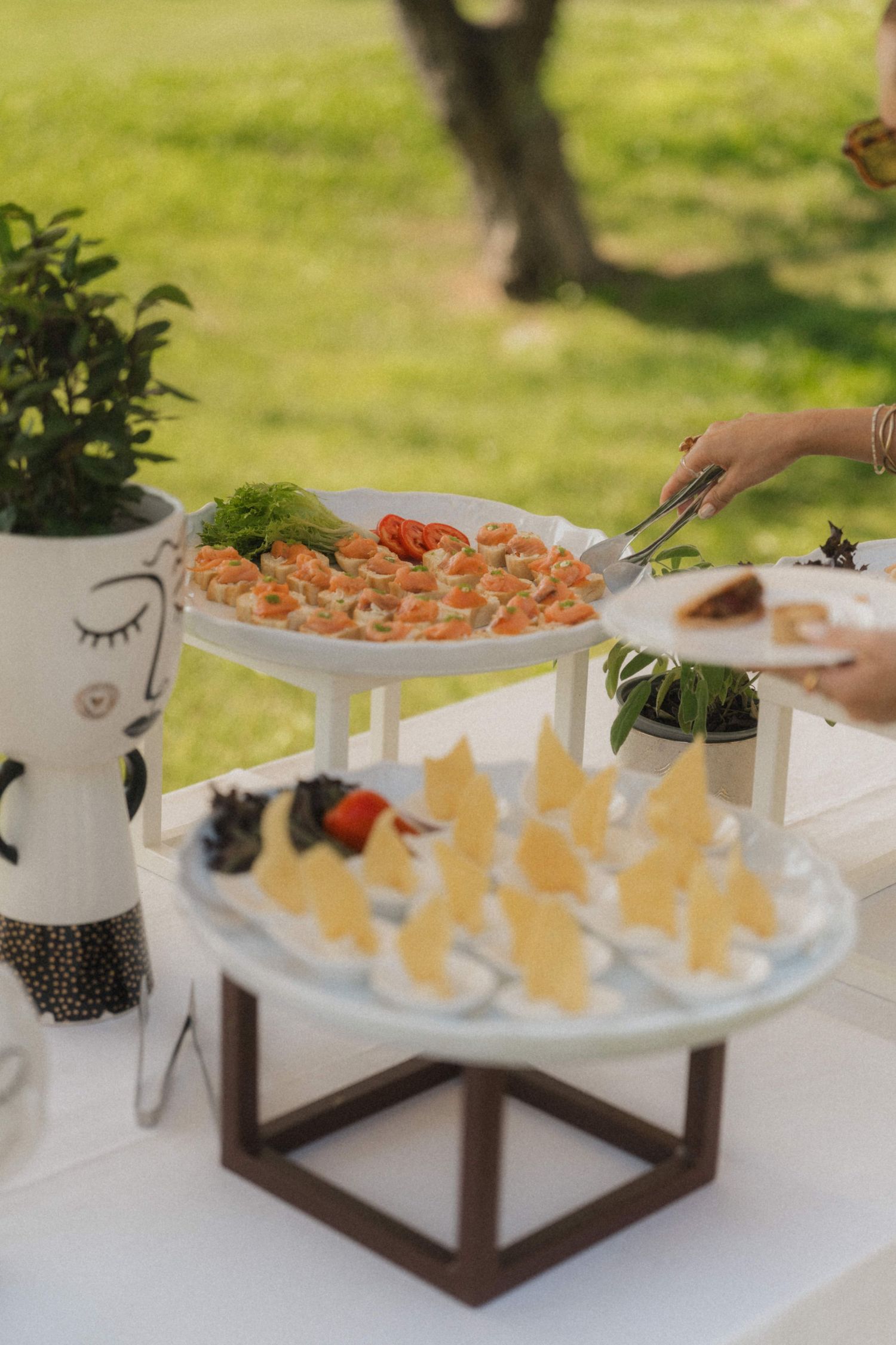 Guest using tongs to serve shrimp appetizers from a tiered white display stand at an outdoor wedding reception.
