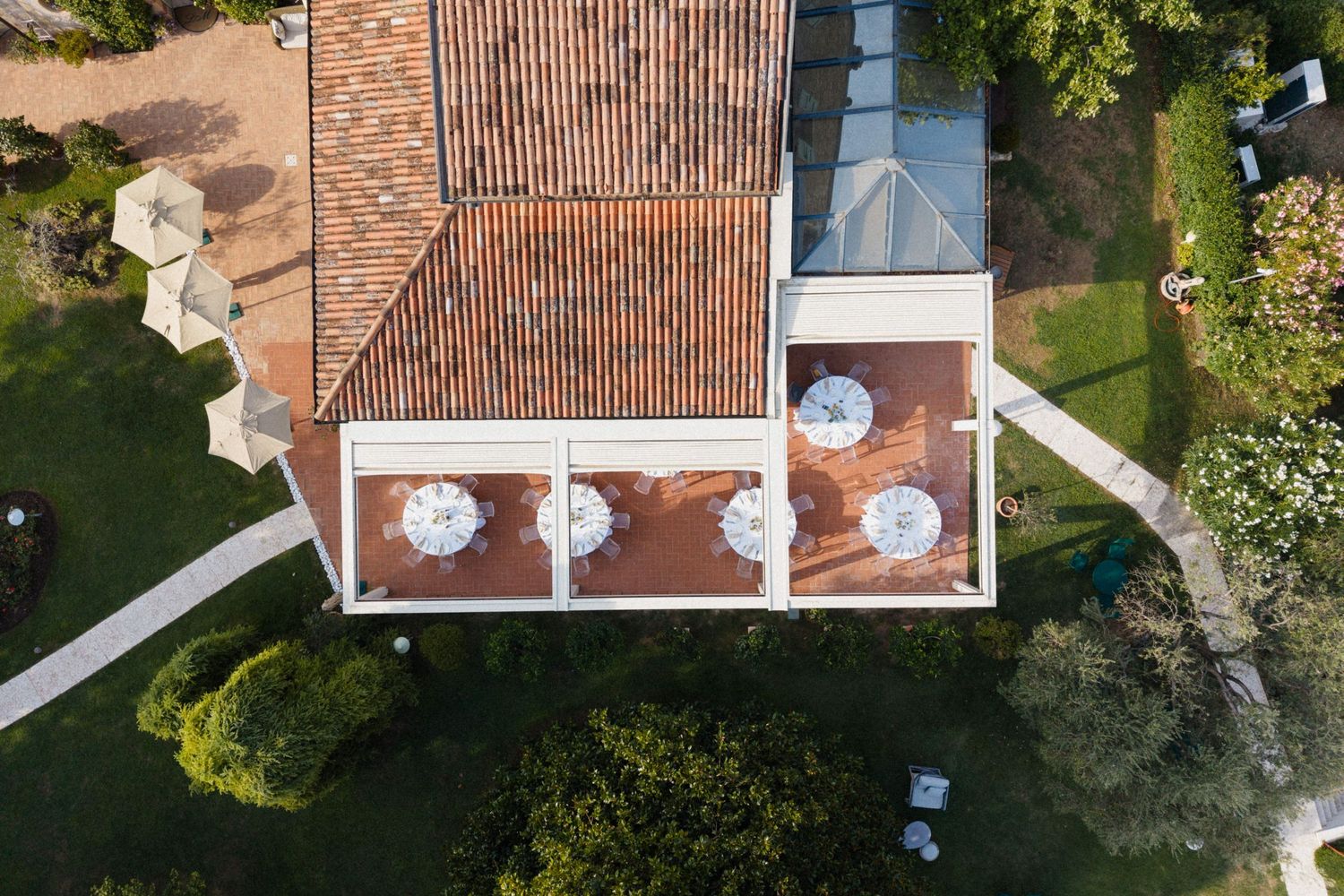Aerial view of a Mediterranean villa with terracotta roof, outdoor terrace with white umbrellas, surrounded by lush gardens.