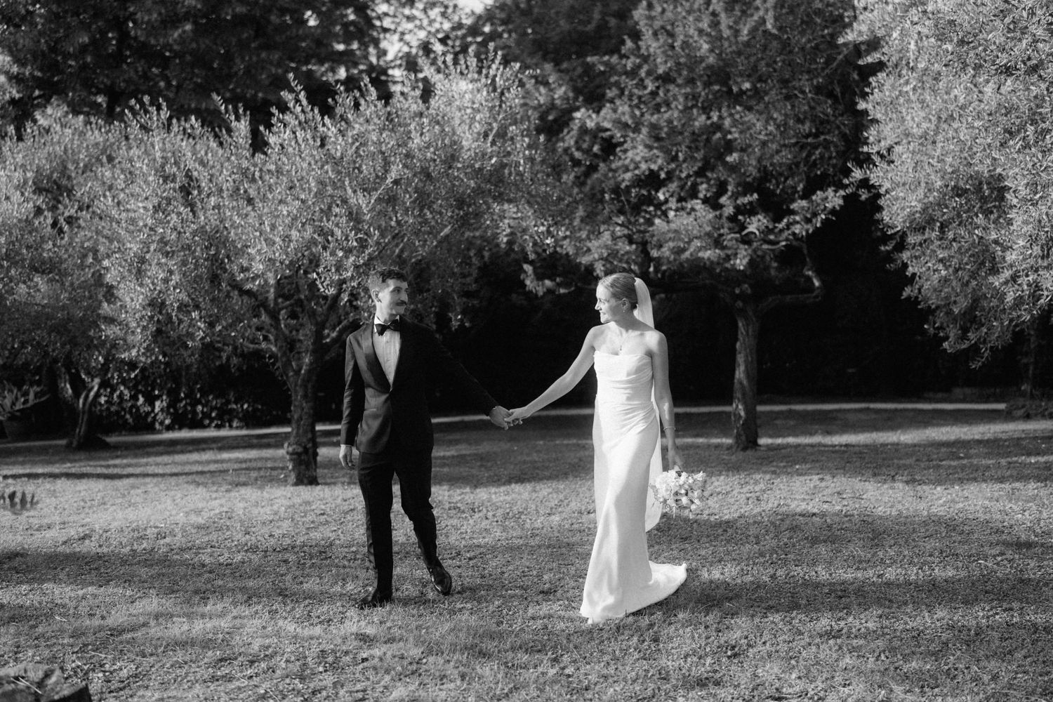 Bride and groom hold hands in a sunlit garden in a black and white wedding photo.