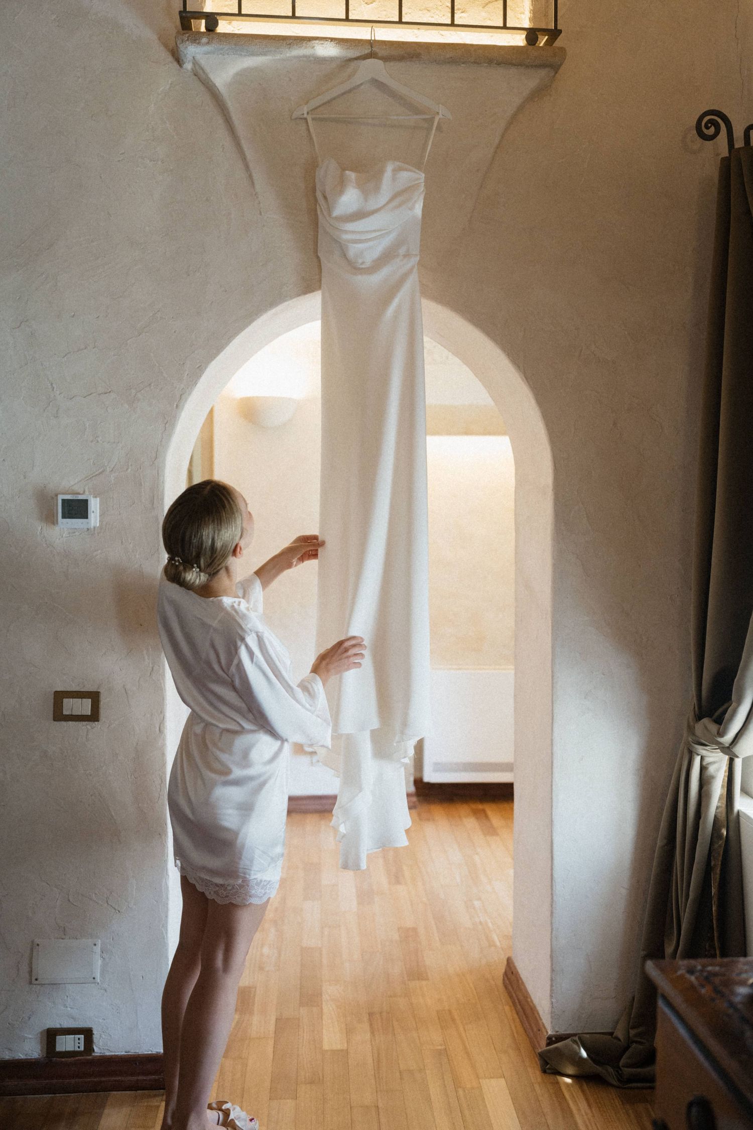 Bride in white robe admiring her elegant wedding dress hanging in a sunlit arch doorway of a rustic venue.