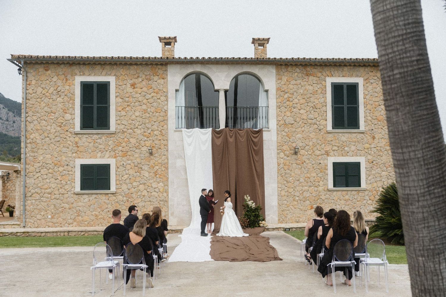 Intimate outdoor wedding ceremony at rustic stone villa with guests seated in ghost chairs.