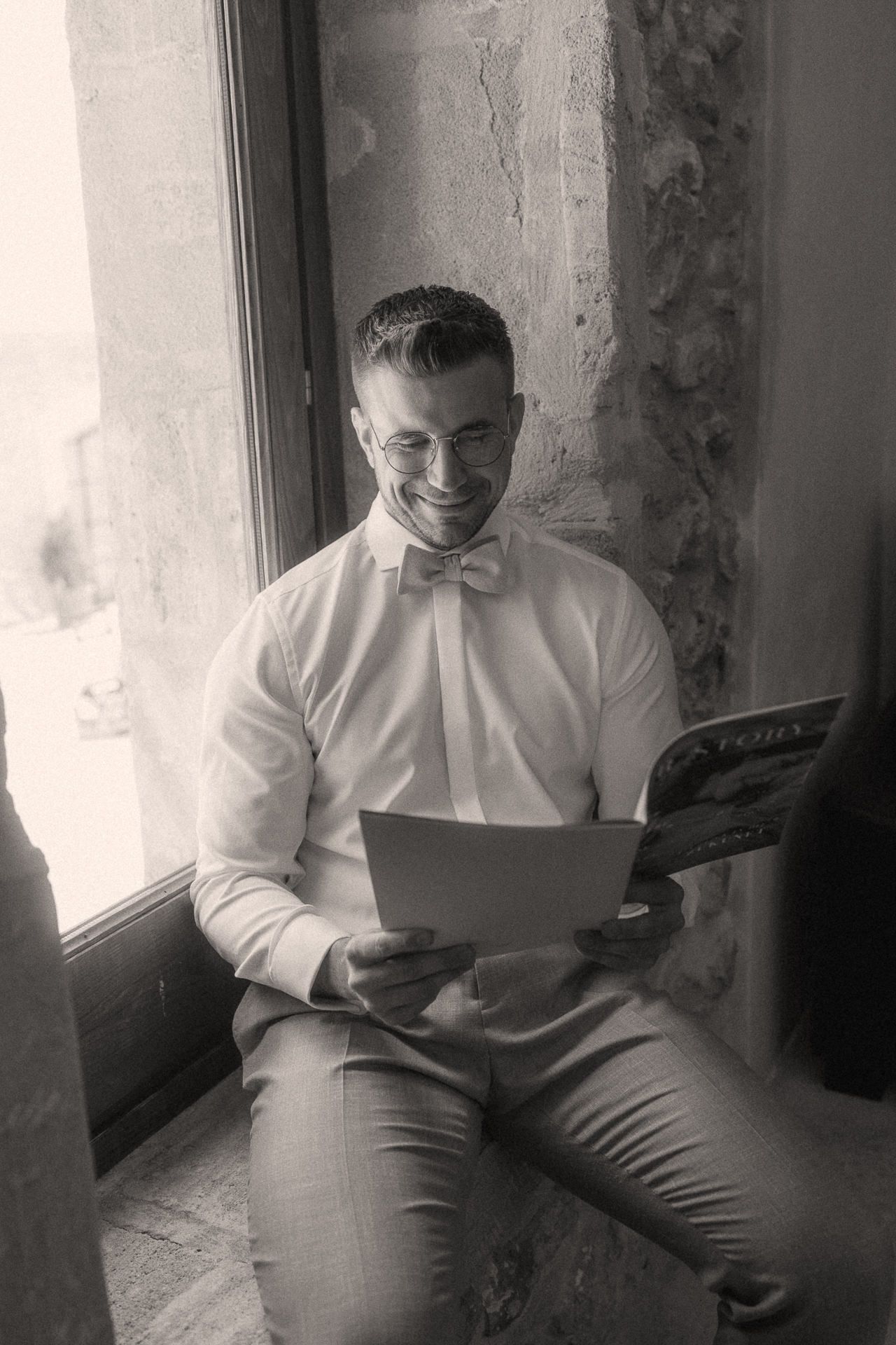 Black and white photo of a smiling groom in white shirt and bow tie reading a card by a window.