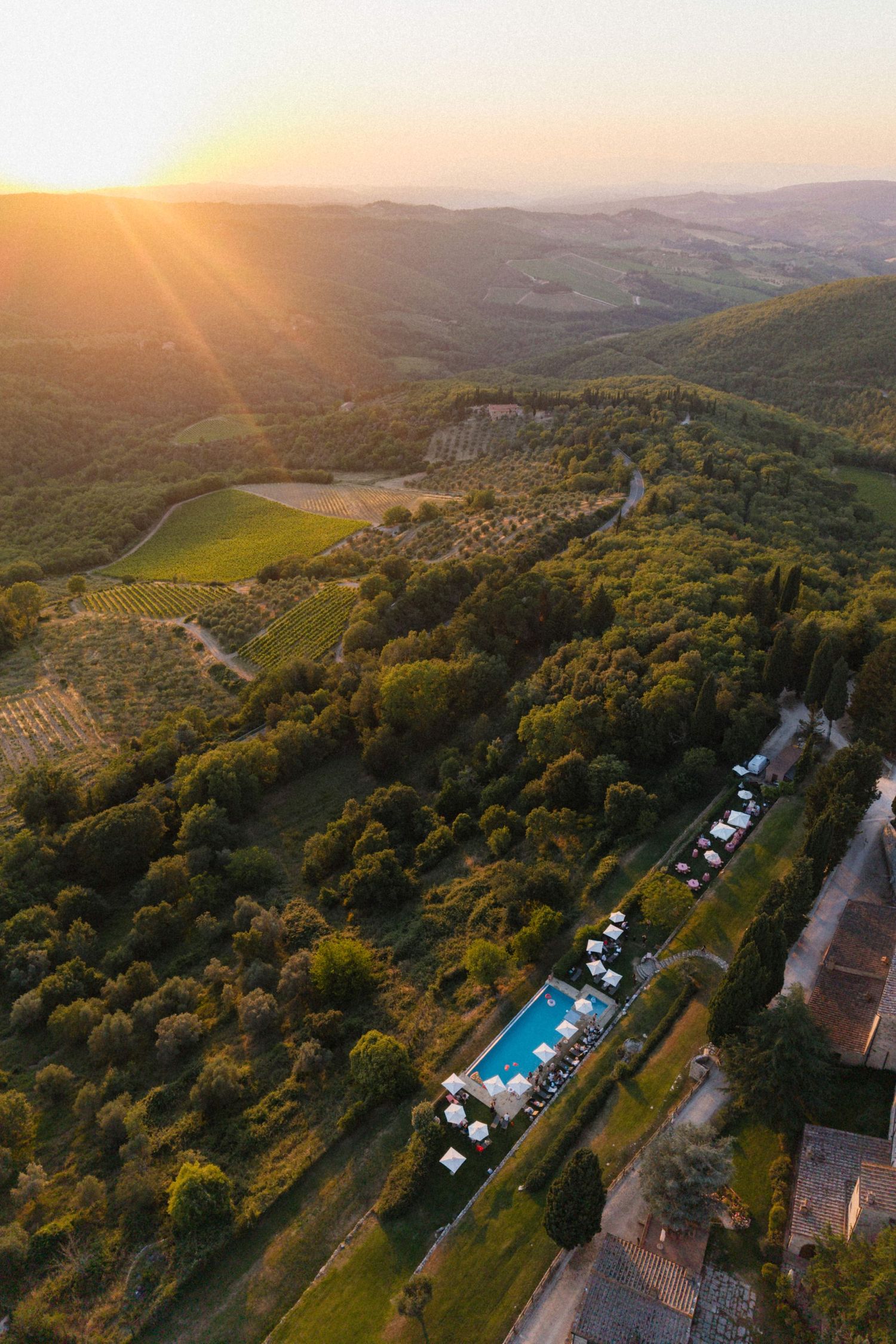 Aerial sunset view of a Tuscan villa with a swimming pool surrounded by vineyards and lush green hills.