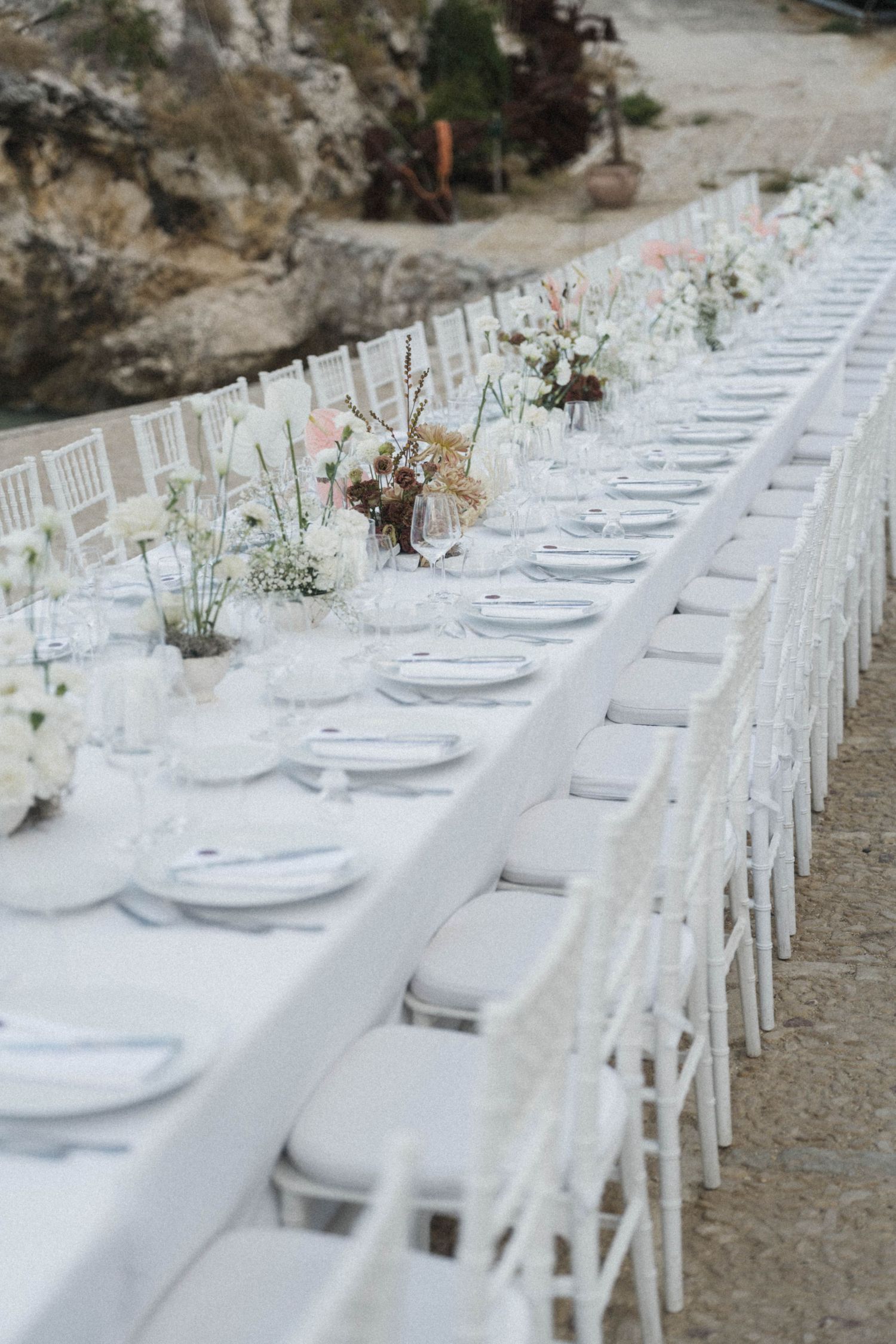 Elegant outdoor wedding reception with long white table, chiavari chairs, and floral centerpieces set against rocky backdrop.