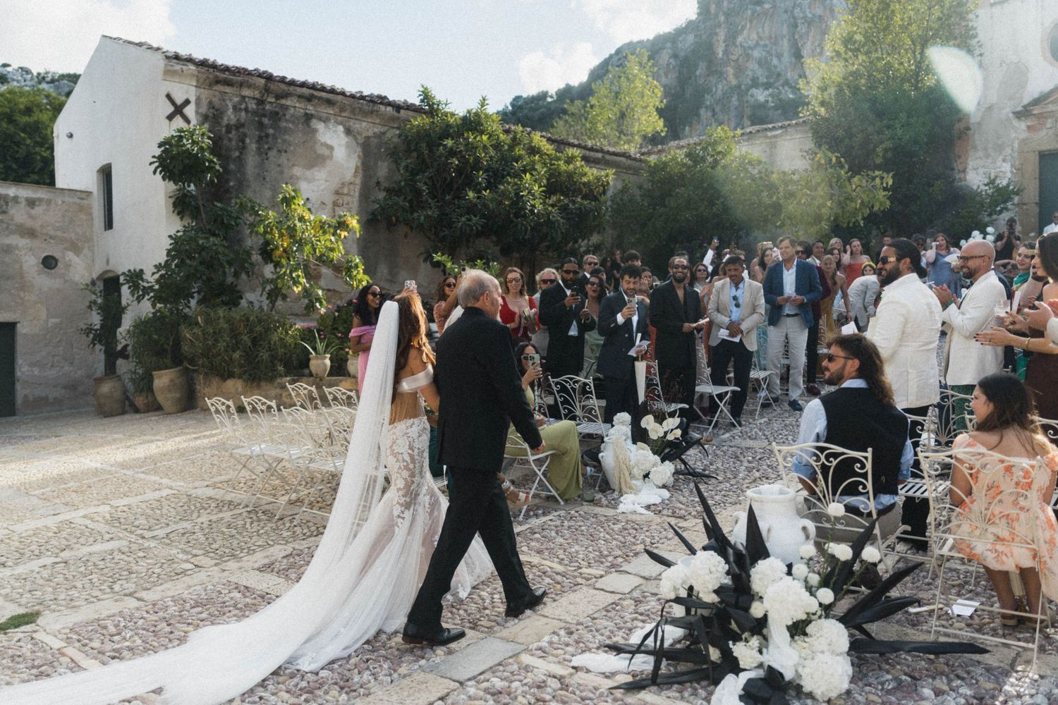 Bride in cathedral veil walks down aisle at rustic outdoor Italian wedding.