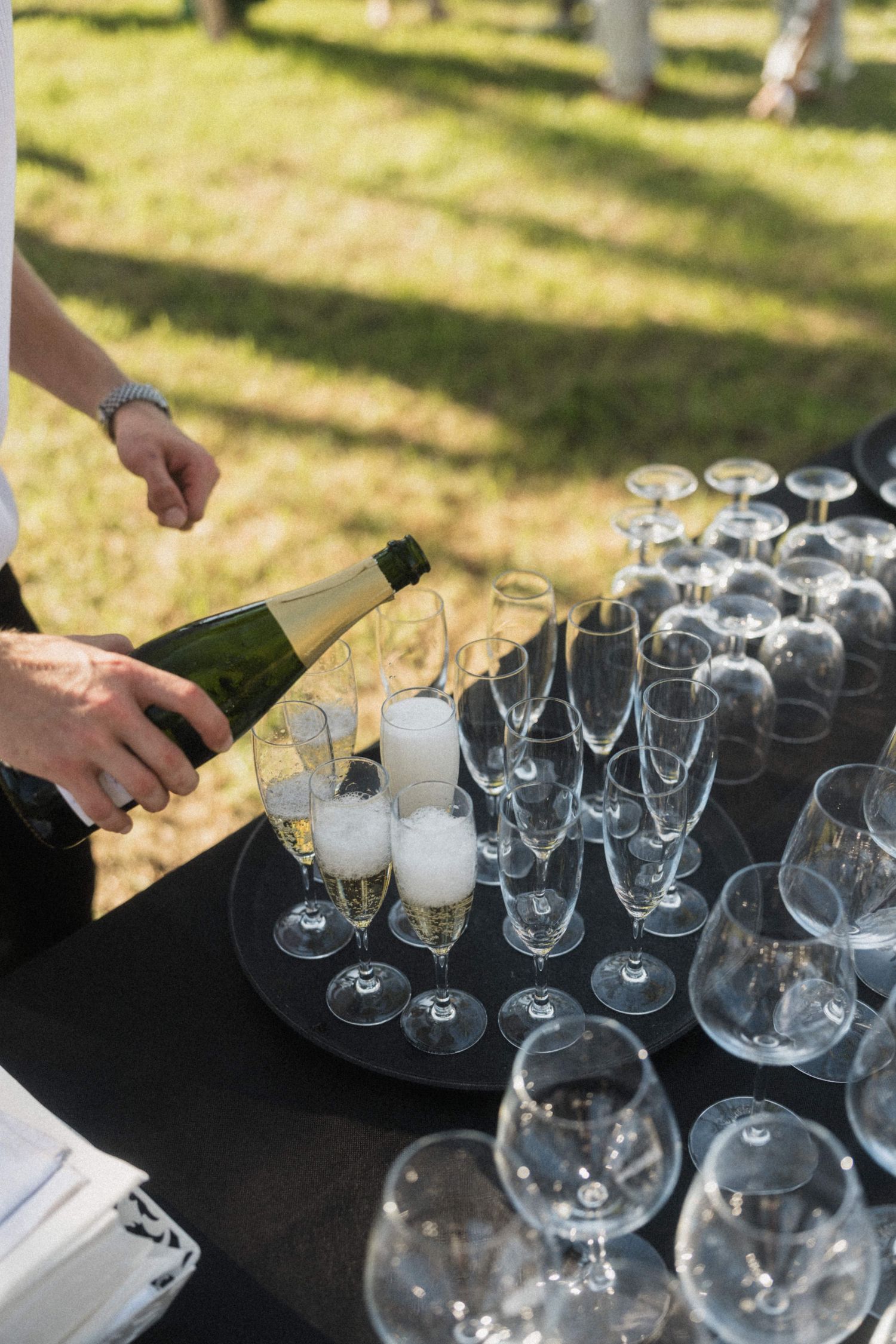 Waiter pouring champagne into flutes at outdoor wedding reception bar setup.