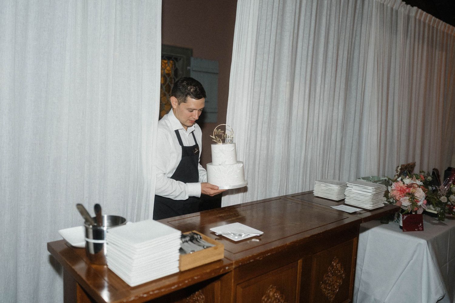 Wedding caterer carefully arranges a white tiered cake on an antique wooden table indoors.
