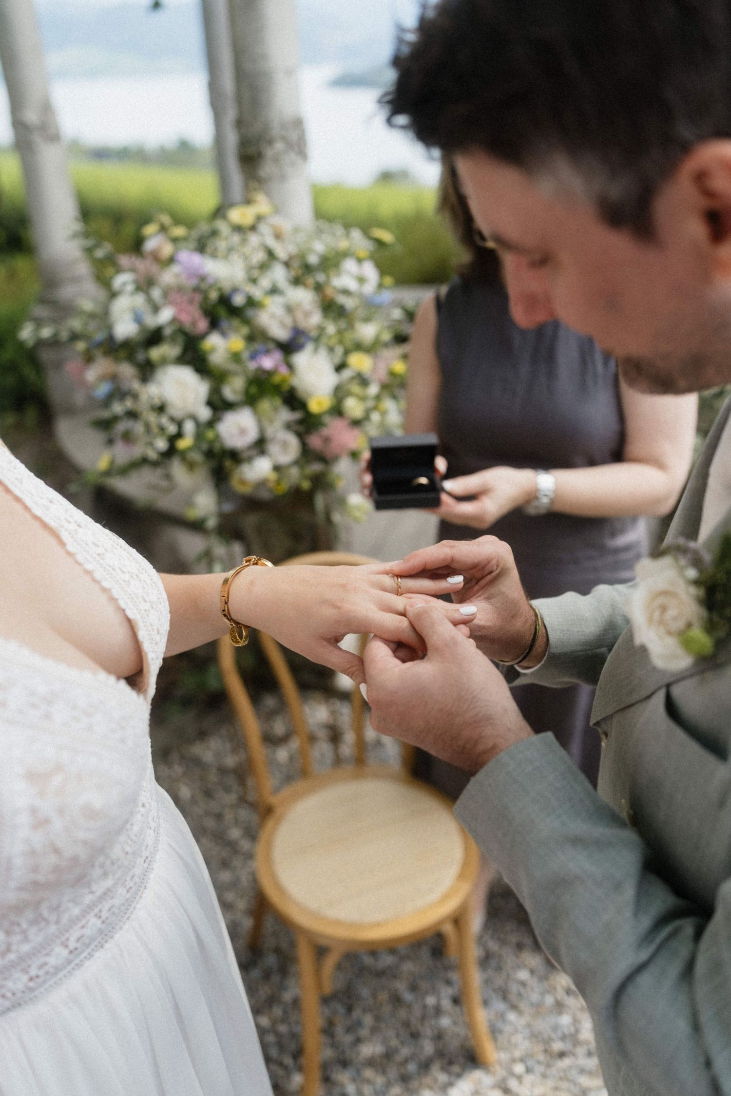 Groom placing wedding ring on brides finger during an outdoor ceremony with floral arrangements in the background.