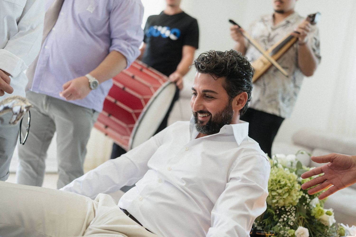 Man in white shirt sits smiling as musicians play drums at a celebratory gathering indoors.