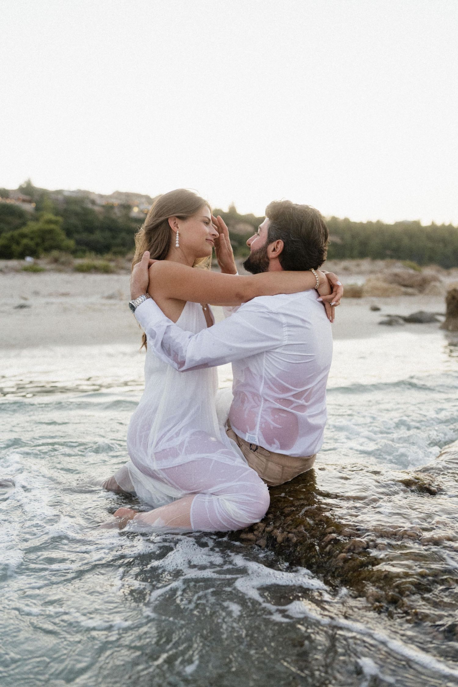 Bride and groom in wet white clothes sit romantically in ocean waves on a rocky beach shore.