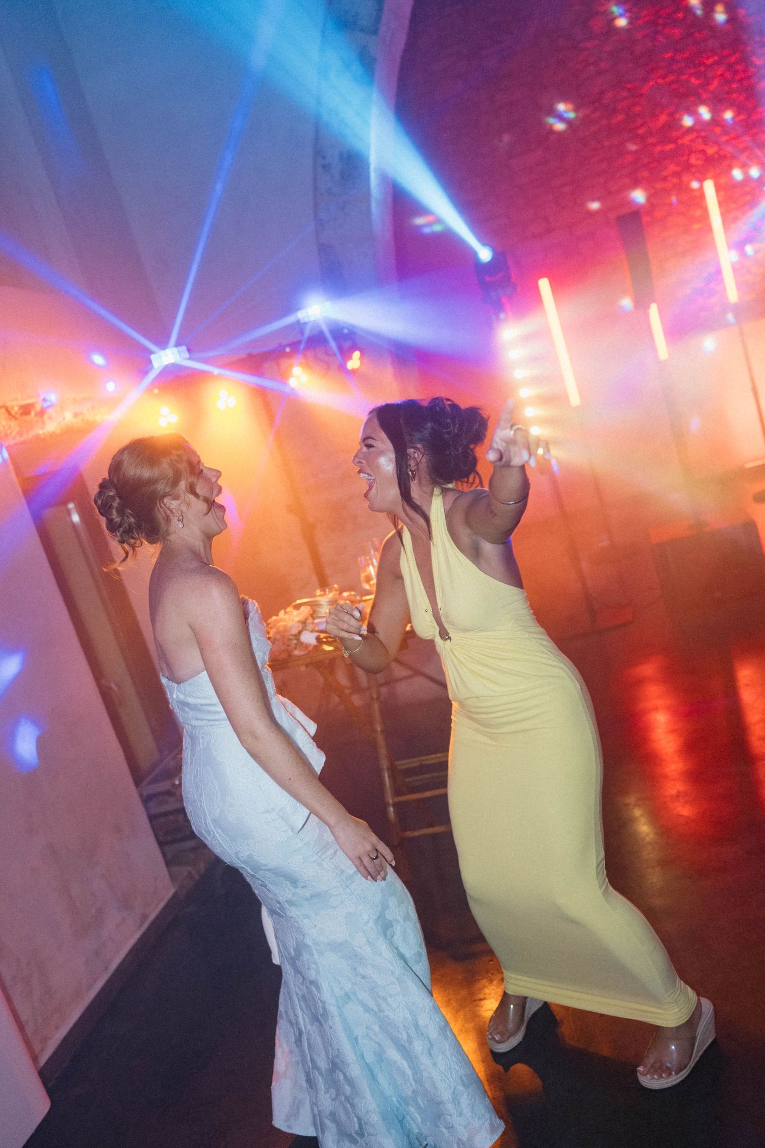 Two women in elegant dresses dancing joyfully at a wedding reception with colorful stage lighting.