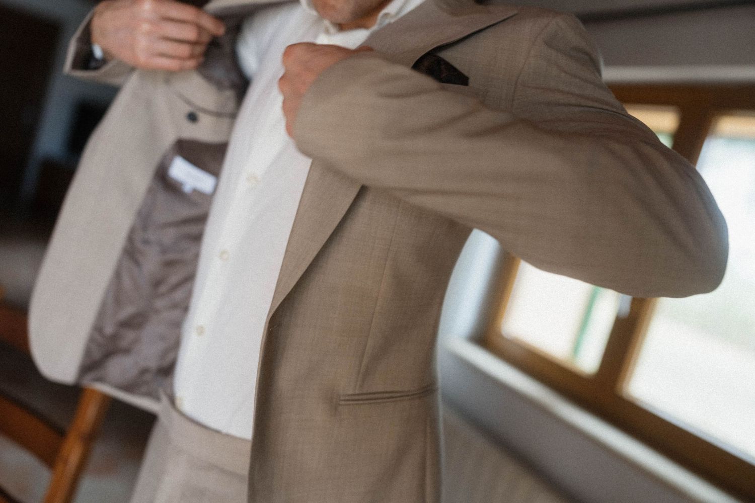 Man in beige suit jacket being helped to dress, adjusting collar indoors.