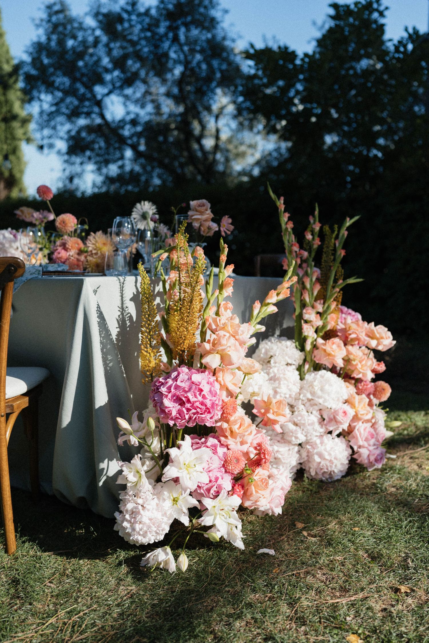 Lush floral arrangement of pink hydrangeas, gladioli, and white lilies cascading beside an outdoor wedding reception table.